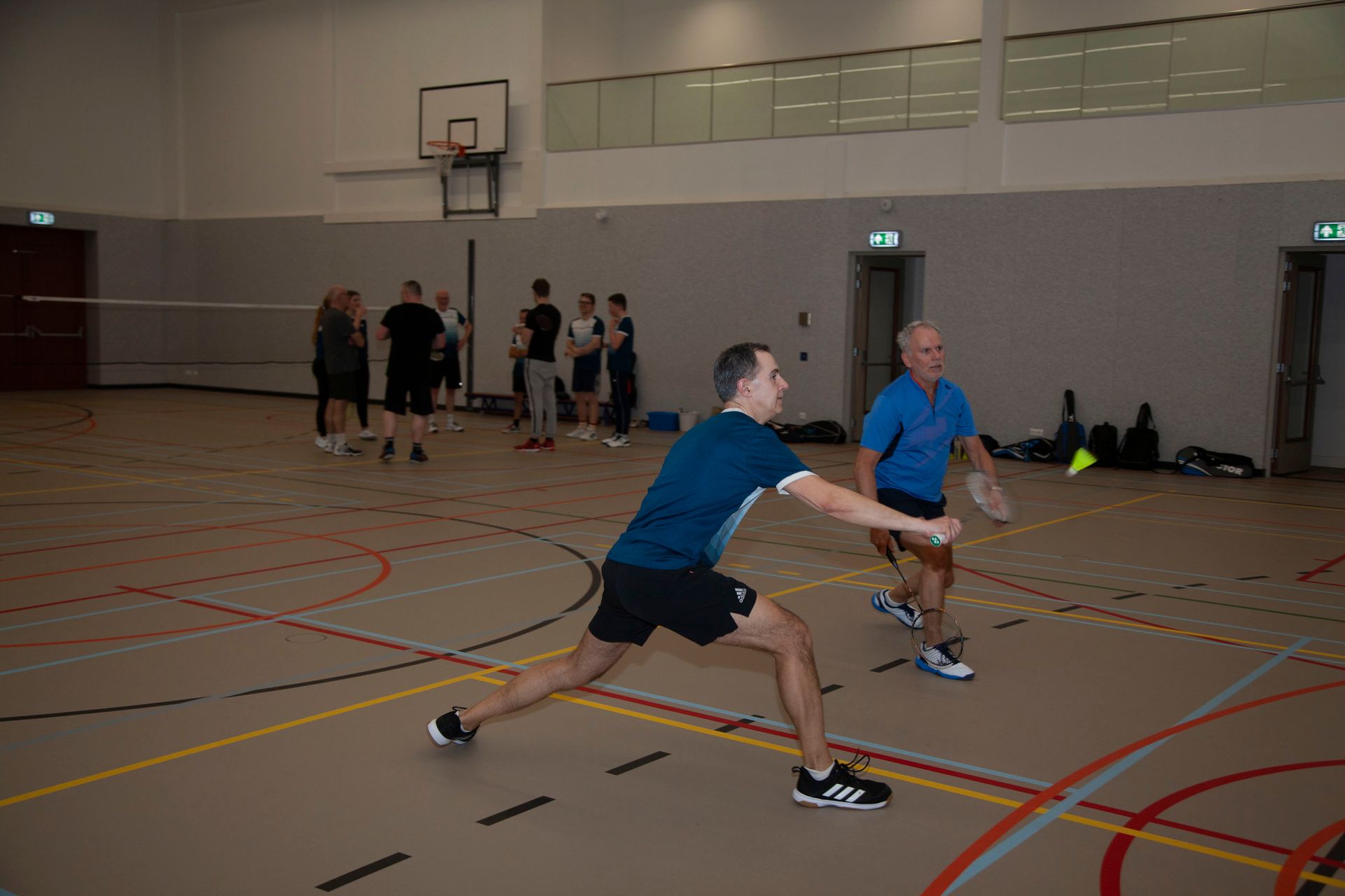 Twee mensen in sportkleding maken lunges op de vloer van een sportschool, met een groep mensen op de achtergrond.