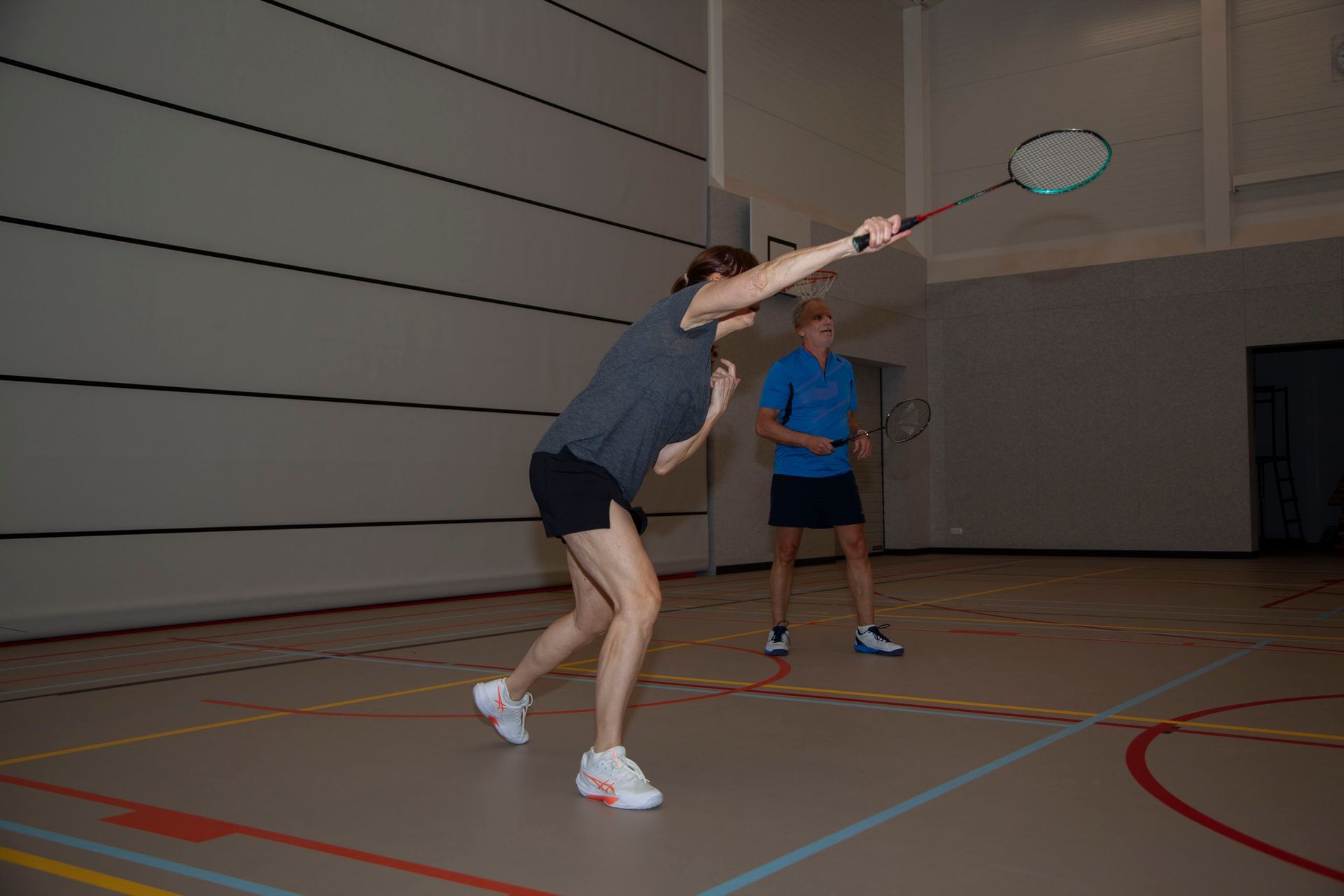Twee mensen spelen badminton op een indoorbaan, waarbij één persoon op de voorgrond zijn racket probeert te zwaaien.