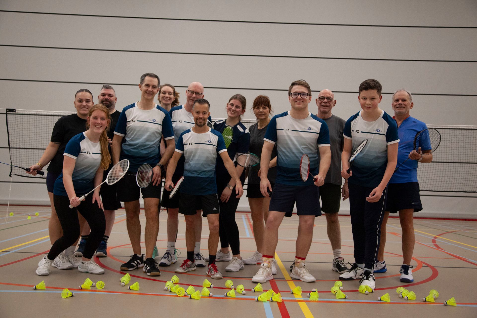 Een groep badmintonspelers in blauw-witte shirts poseert voor een foto op een baan waar shuttlecocks op de grond liggen.