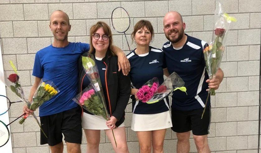 Vier mensen staan lachend voor een bakstenen muur, met boeketten bloemen in hun handen en gekleed in sportkleding.