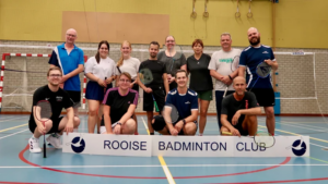 Een groep van twaalf badmintonspelers in sportkleding poseert achter een spandoek van de Rooise Badminton Club