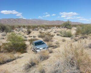 Damaged silver car off-road in desert, mountains in background, under blue sky.