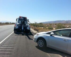 A tow truck pulling a silver Prius off the side of a desert highway under a blue sky.