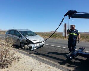 A silver Prius with front-end damage being towed by a tow truck driver on a desert highway.
