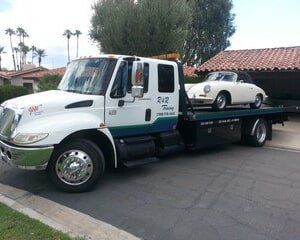 A white tow truck with a vintage white Porsche on the flatbed, parked on a driveway.