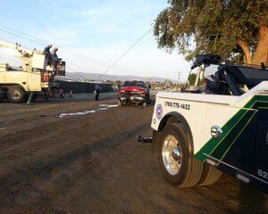 Tow truck pulling a red pickup truck, power lines overhead, workers on a boom lift, dirt road setting.