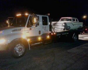 Tow truck at night carrying a vintage white car; headlights and lights illuminate.