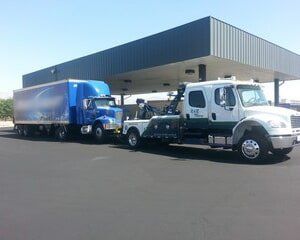 White tow truck towing a blue semi-truck at a gas station, under a canopy, on a sunny day.