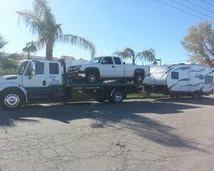 Tow truck transporting a white pickup truck and a camping trailer on a sunny day.