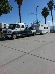 A white tow truck towing a white travel trailer on a paved surface under a blue sky.