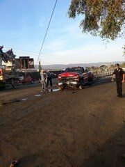Red pickup truck damaged, people assessing the damage. Dirt road, utility poles visible.