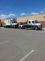 A white tow truck towing a white refrigerated semi-truck on a paved surface. Blue sky and building in background.