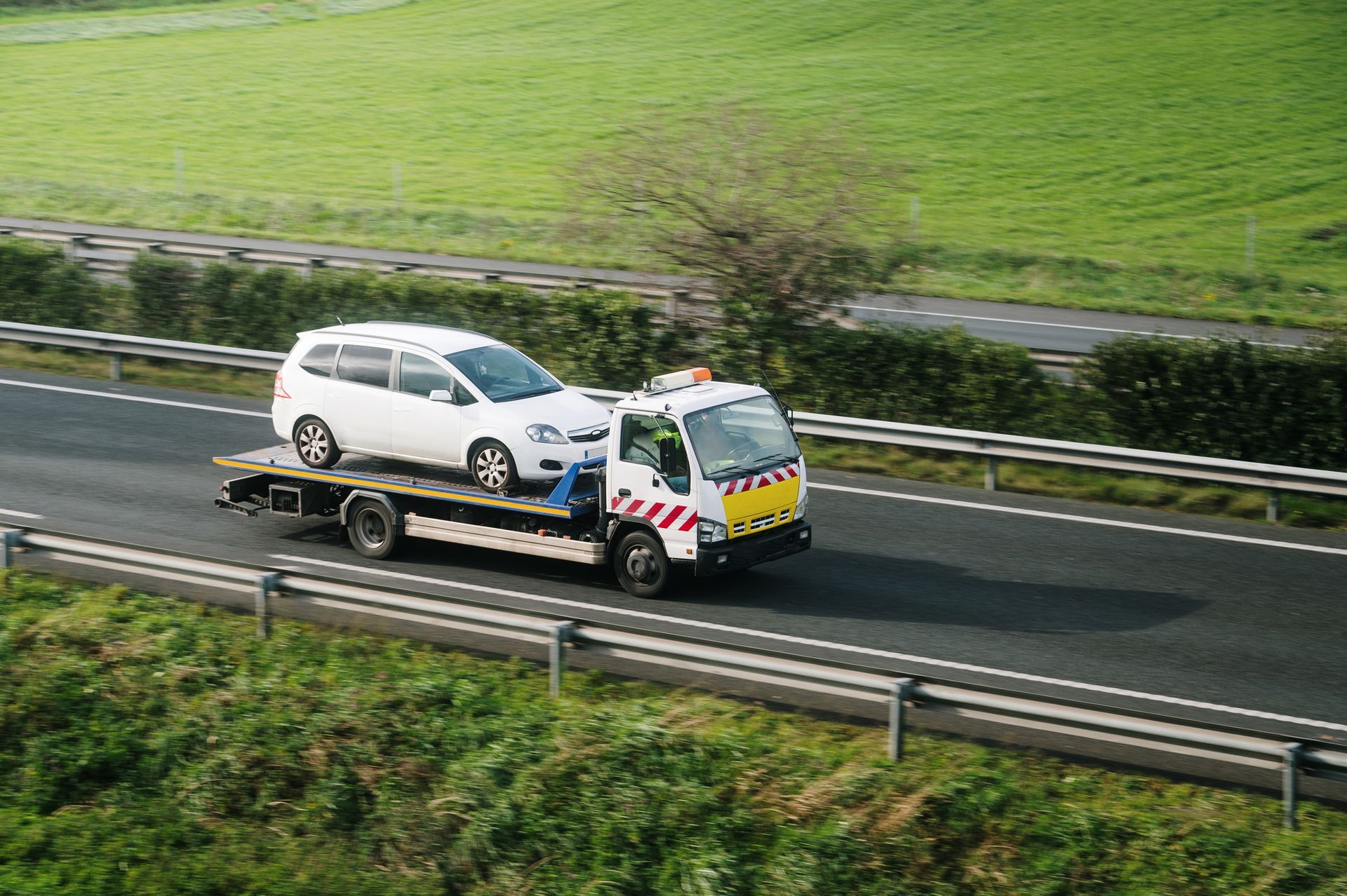 White minivan being towed on a flatbed tow truck on a highway. Green fields in background.