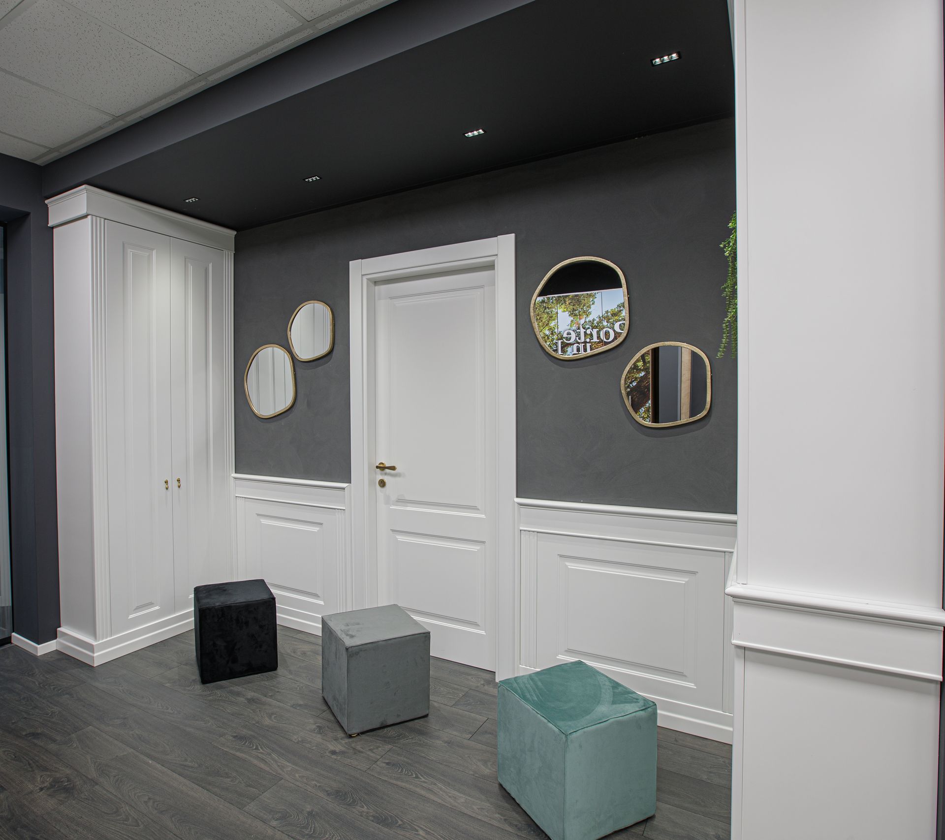 Un corridoio moderno con finiture bianche, pareti grigio scuro, tre pouf cubici e specchi decorativi.