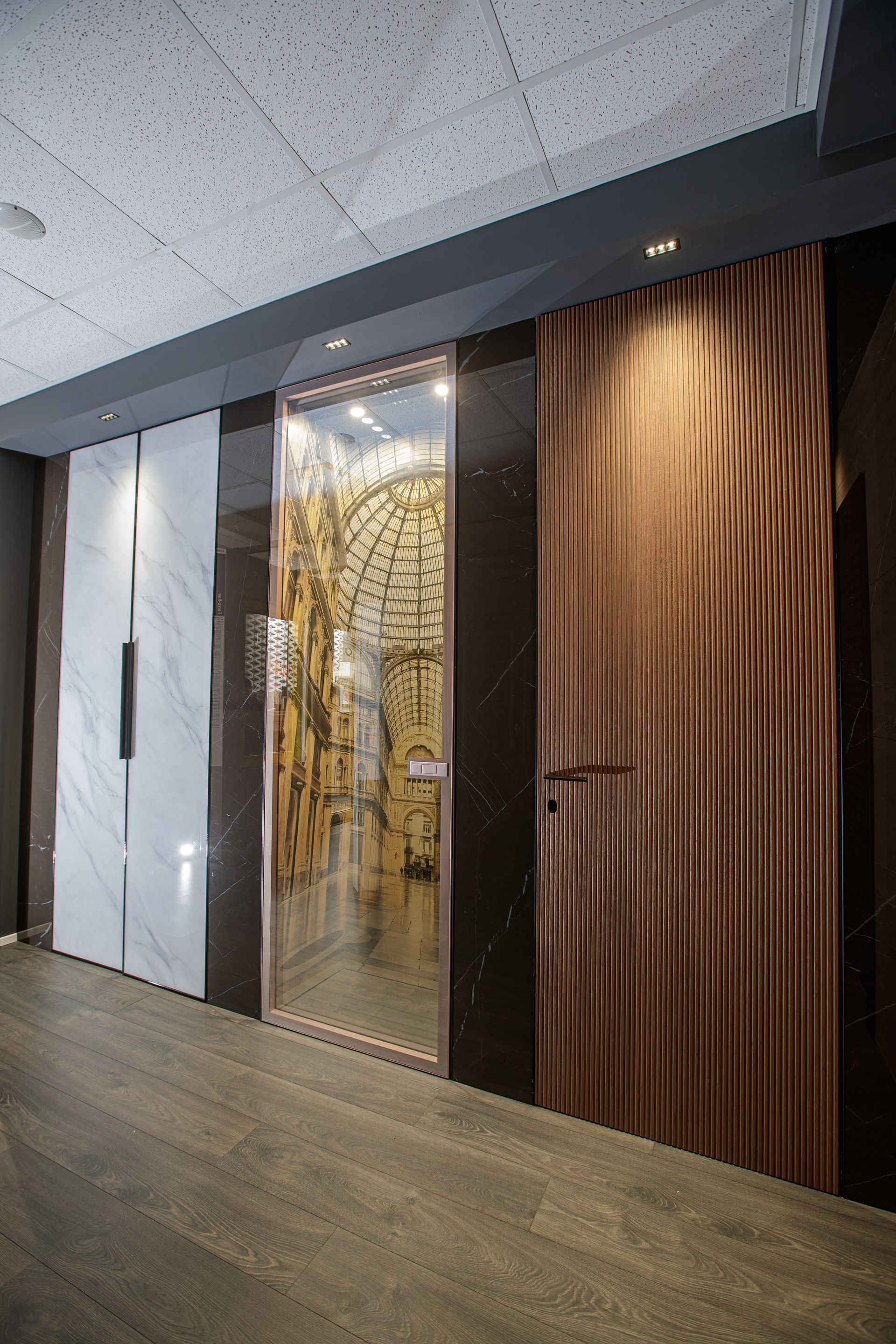 Corridoio con tre pannelli murali testurizzati: marmo bianco, porta in vetro con dettagli dorati e rame ondulato.