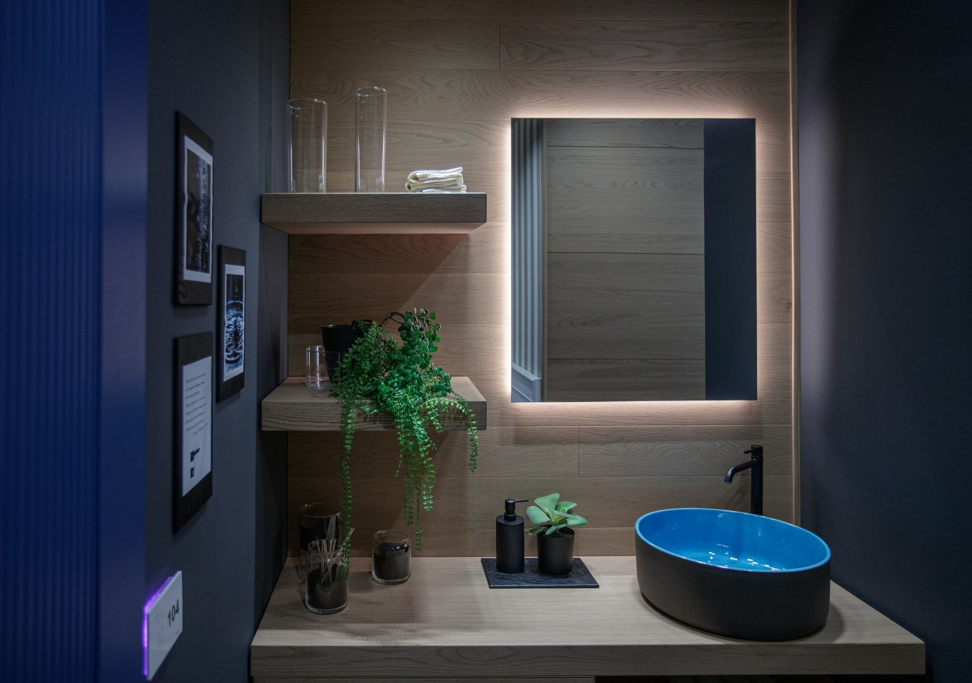 Bagno moderno con specchio retroilluminato, lavandino blu, ripiani in legno e pareti scure.