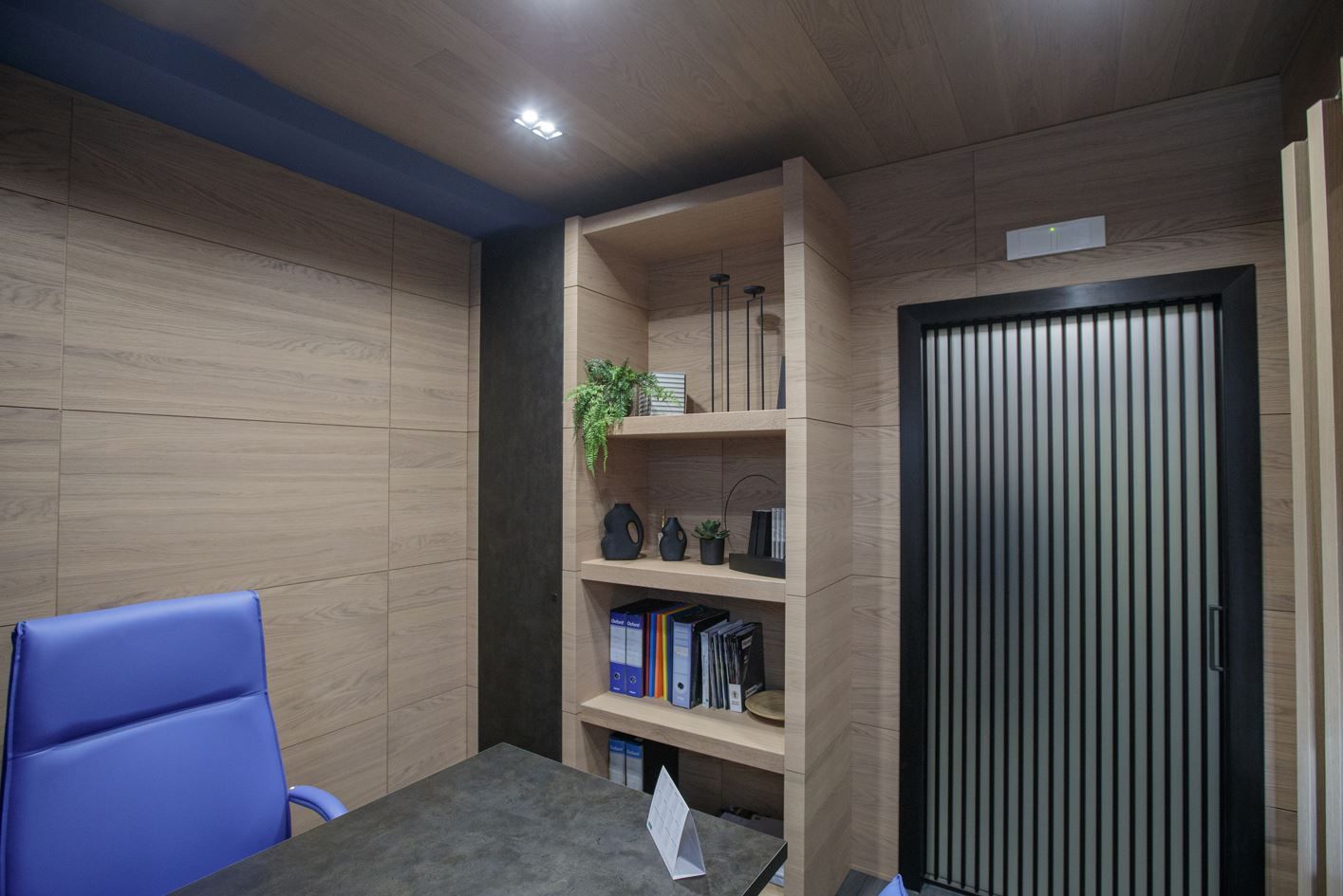 Interno dell'ufficio: pannelli in legno chiaro, scaffali integrati con oggetti, sedia blu, porta nera.