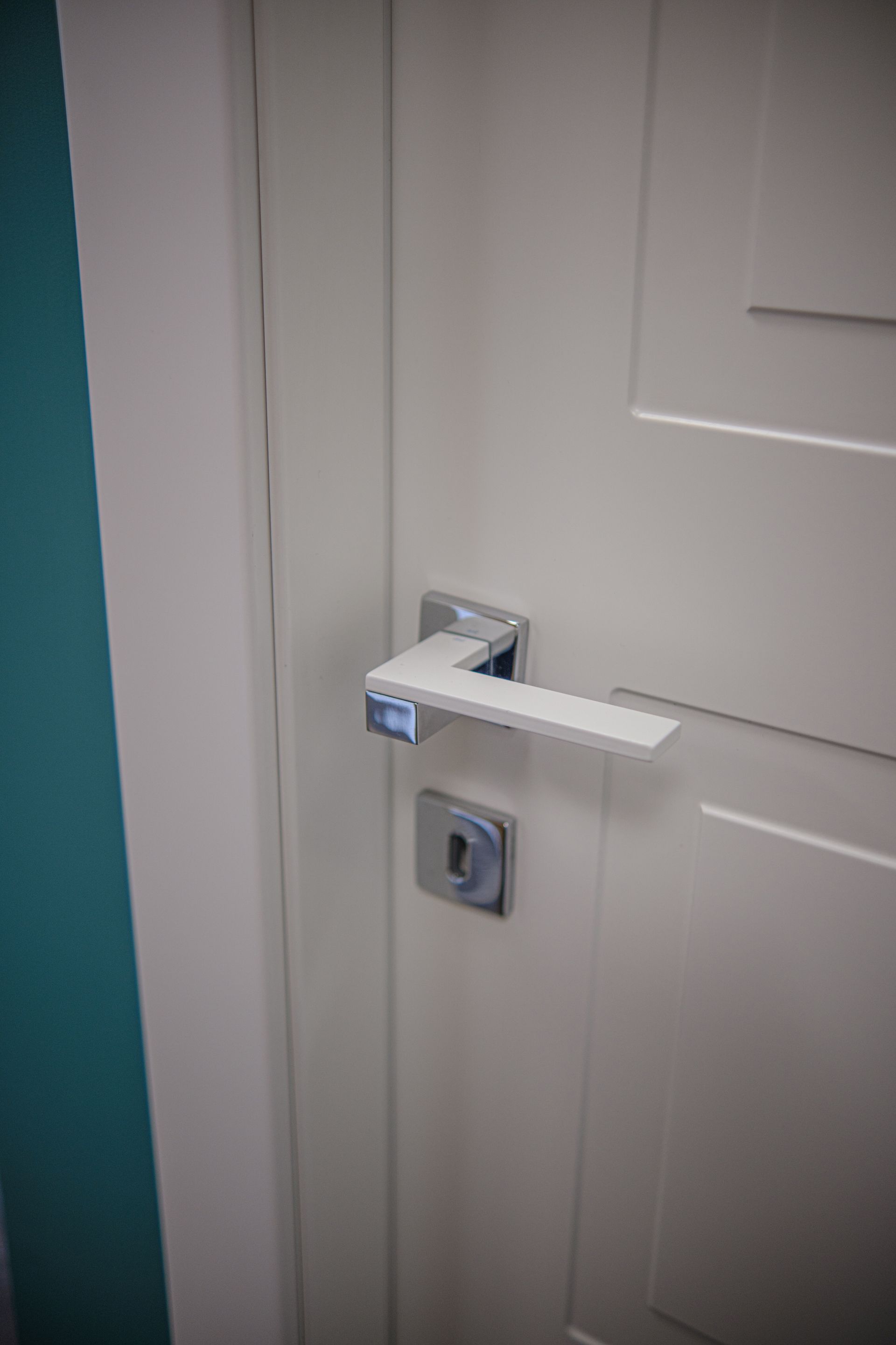 Porta bianca con maniglia e serratura argento e bianche. Parete verde acqua visibile.