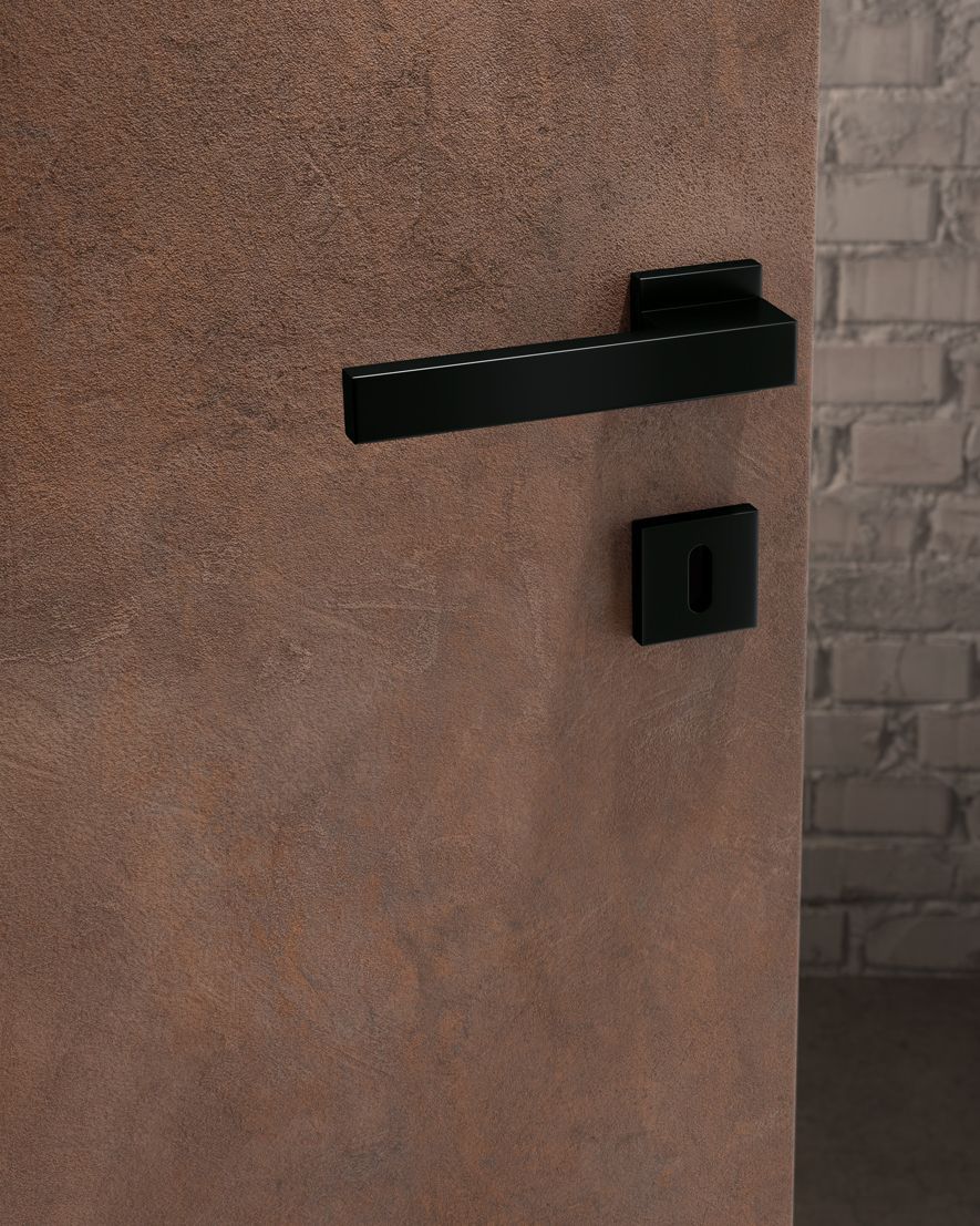 Maniglia e serratura nere su una porta marrone testurizzata, con un muro di mattoni sullo sfondo.