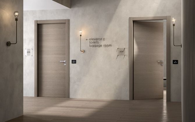 Corridoio con due porte in legno, cartelli direzionali e luci a parete.