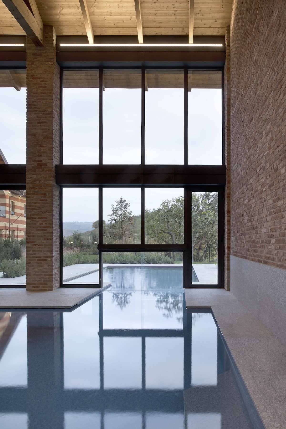 Piscina coperta con ampie vetrate che si affacciano sugli alberi. Pareti in mattoni e travi in ​​legno incorniciano la vista.