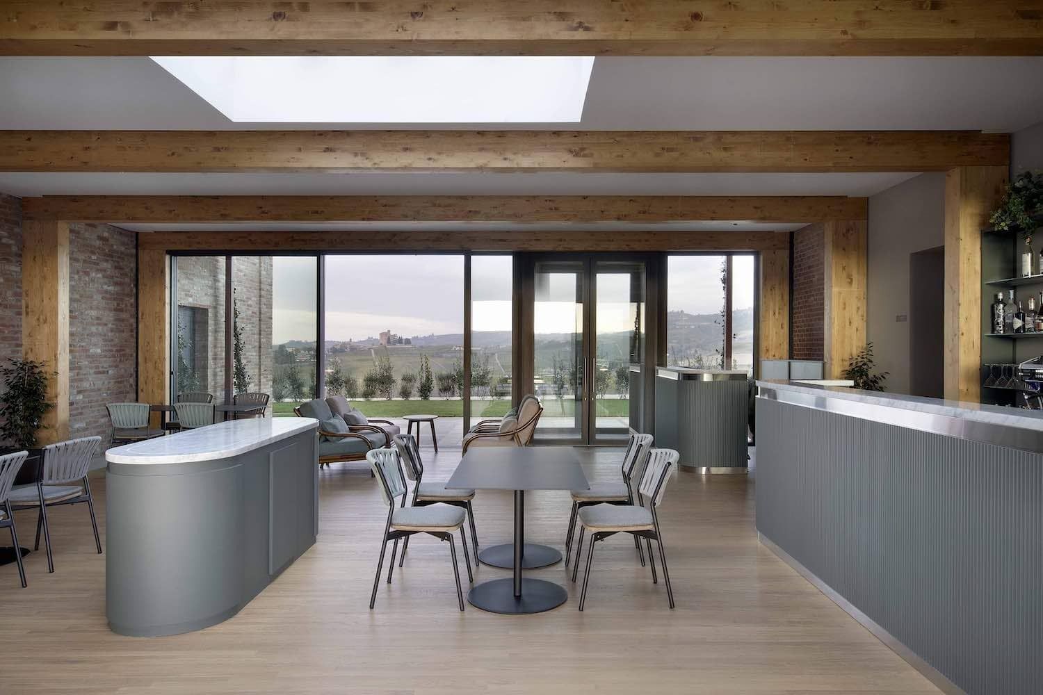 Interno di un bar moderno con ampie finestre, travi in ​​legno, mobili grigi e un tavolo con sedie.