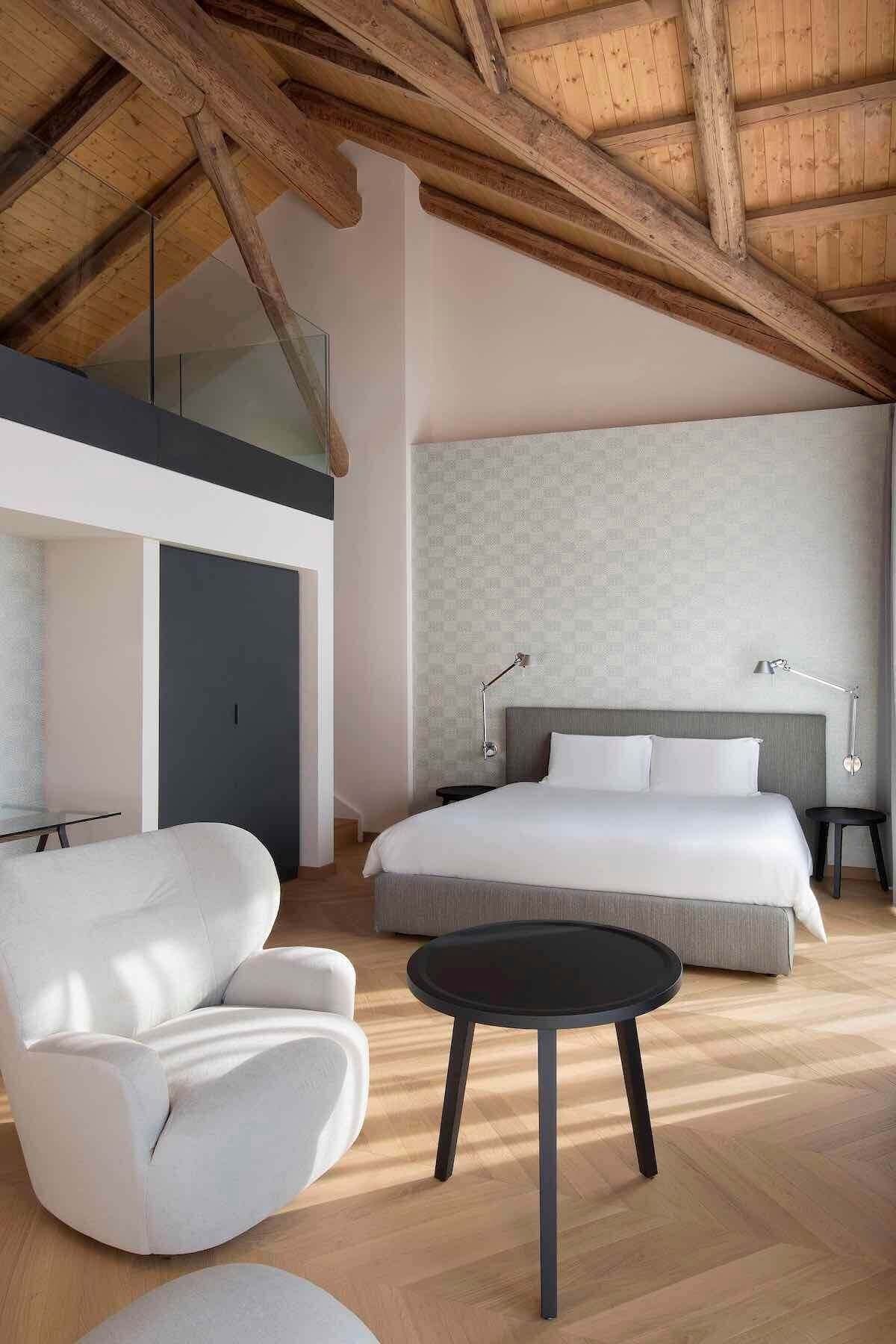 Camera da letto con travi in ​​legno a vista, letto, sedia bianca e tavolino nero.