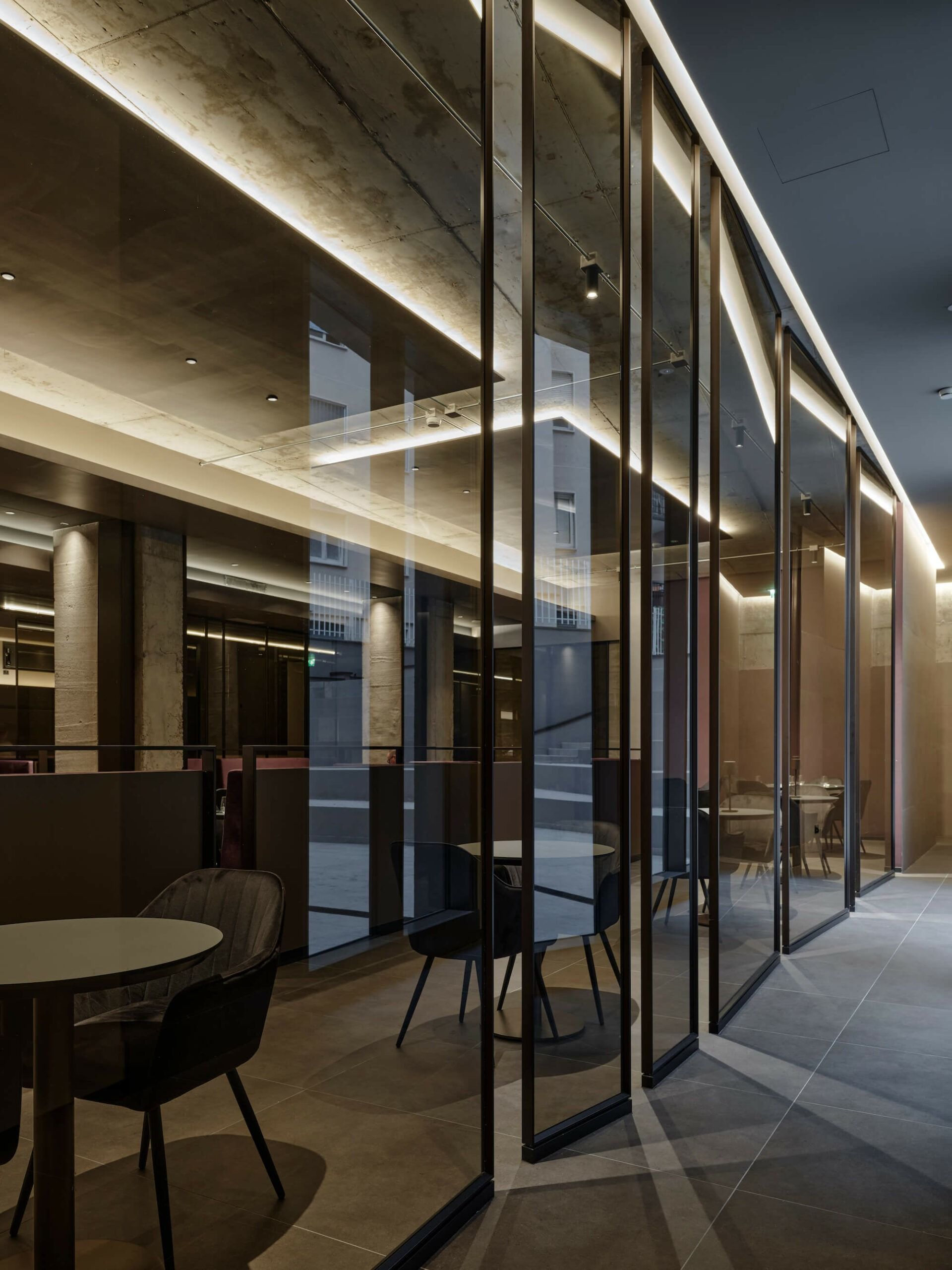 Interno del bar con pareti in vetro, tavoli e sedie, cornici scure e illuminazione ambientale, soffitto in cemento.