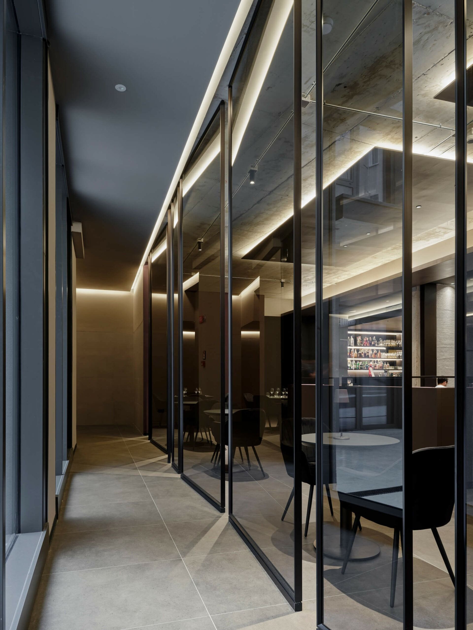 Un corridoio stretto con sale da pranzo con pareti in vetro. Sedie e tavoli neri sono visibili. Luci soffuse.