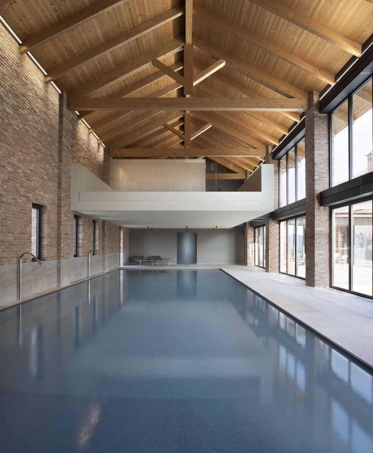 Piscina coperta con travi in ​​legno a vista, pareti in mattoni e ampie finestre.