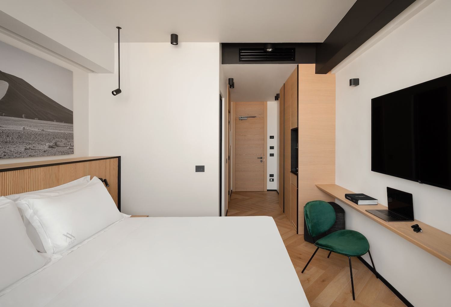 Camera d'albergo con letto, TV e piccola scrivania. Pareti bianche, dettagli in legno e una sedia verde.