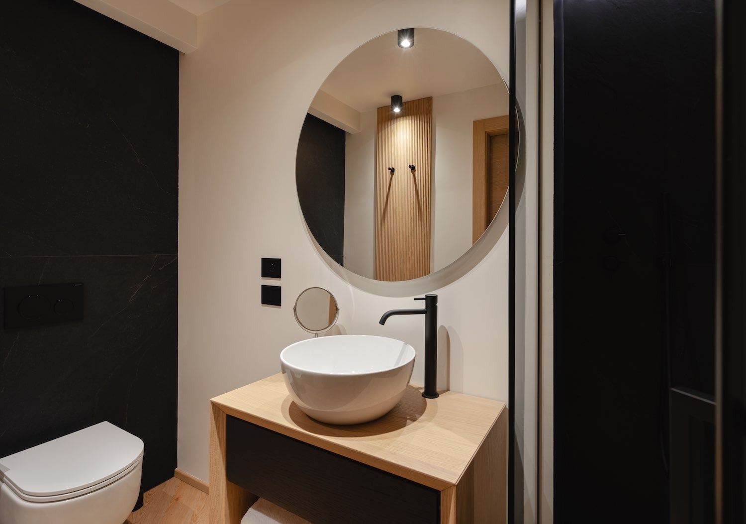 Bagno con specchio rotondo, lavandino bianco, rubinetto nero e WC. Presenta legno e pareti nere.