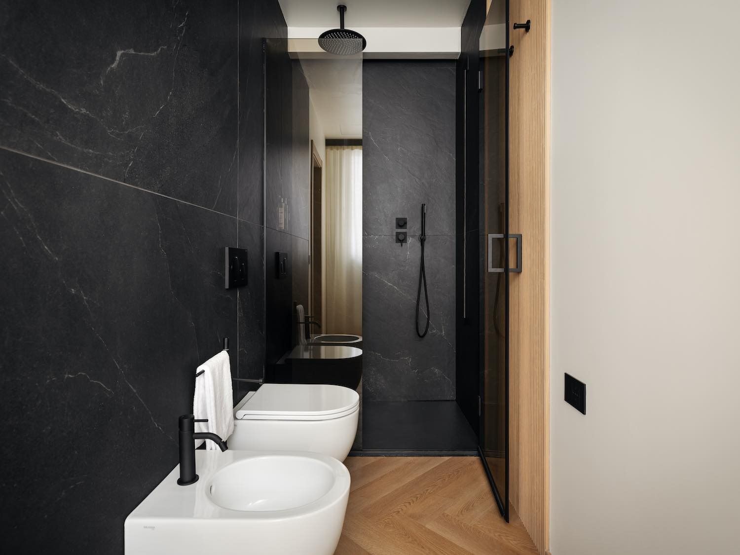 Bagno moderno con pareti in marmo nero, doccia, WC e bidet bianchi; pavimento in legno a spina di pesce.