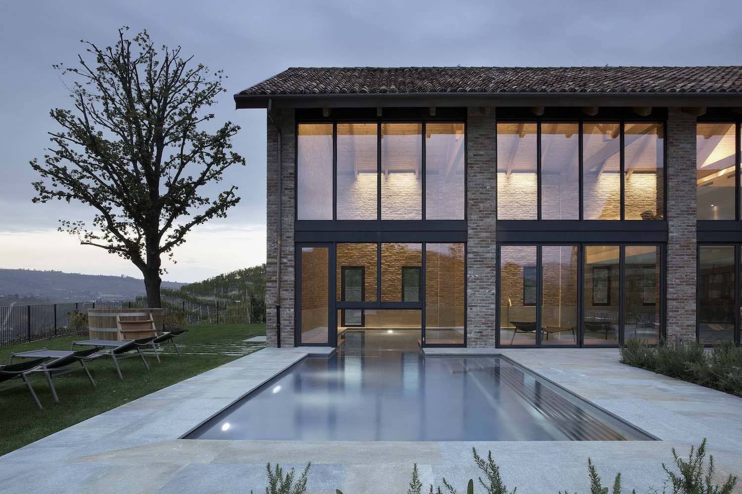 Moderna casa in pietra con ampie finestre, piscina e albero, illuminata al tramonto.