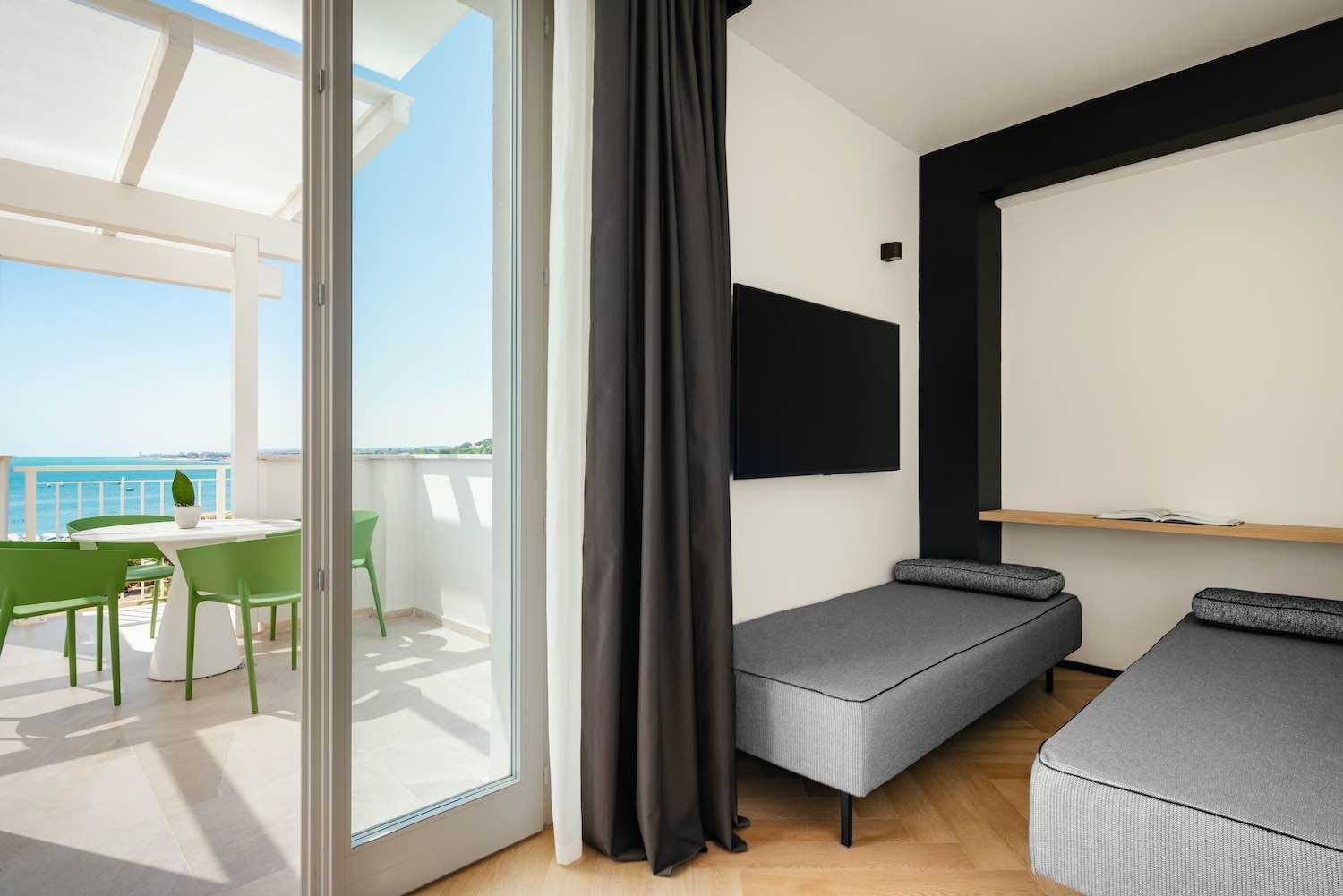 Camera d'albergo con balcone con vista sull'oceano. Due letti singoli, TV, tende grigie e mobili da giardino verdi.