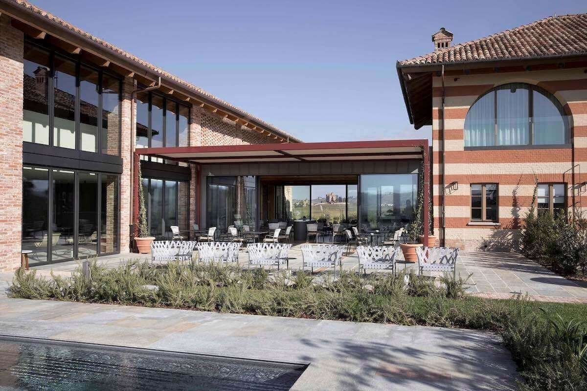 Patio esterno di un edificio in mattoni con ampie finestre. Tavoli e sedie sotto una tettoia, cielo azzurro.