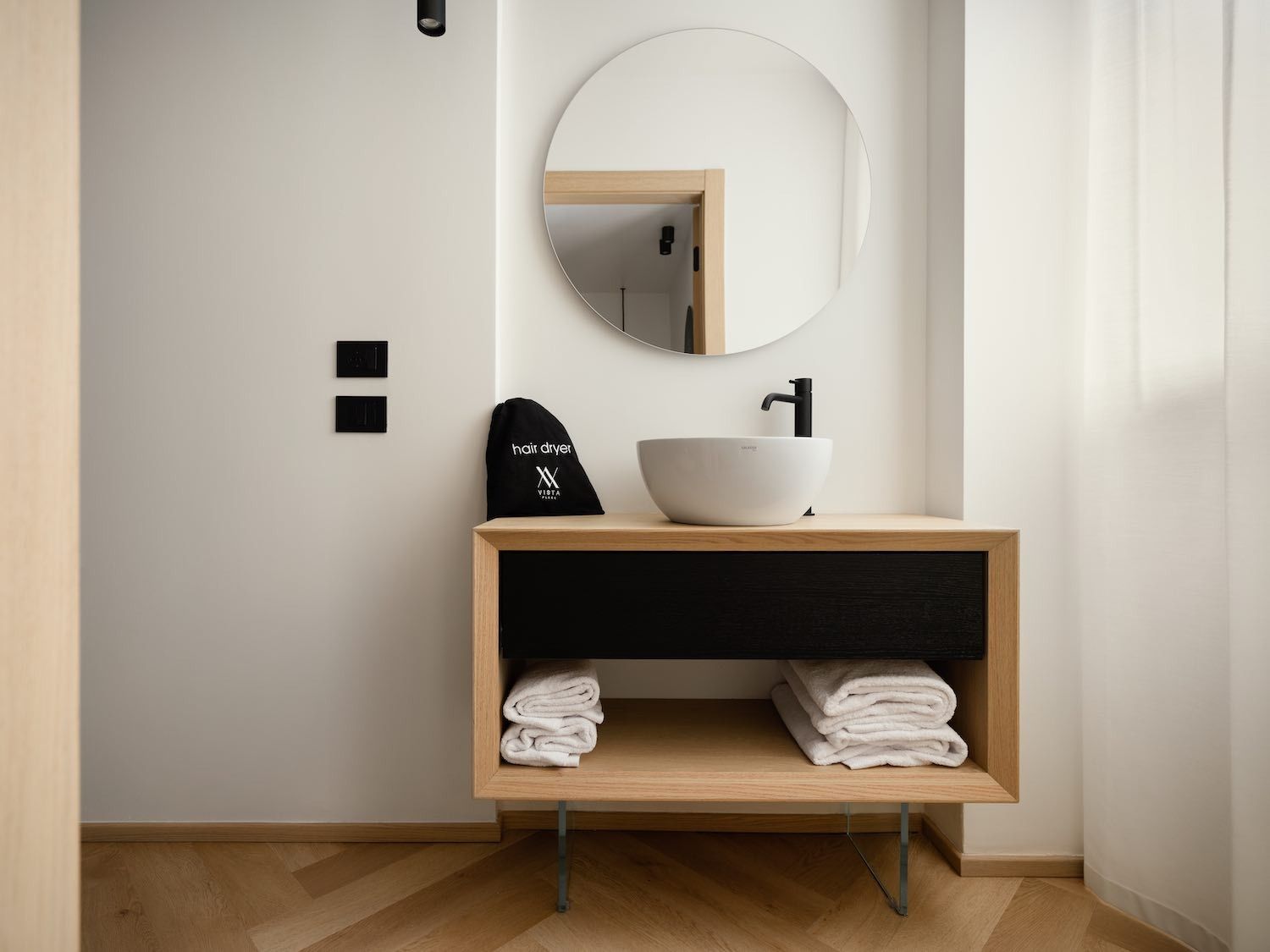 Bagno moderno con specchio rotondo, rubinetto nero e mobiletto nero e in legno con asciugamani.