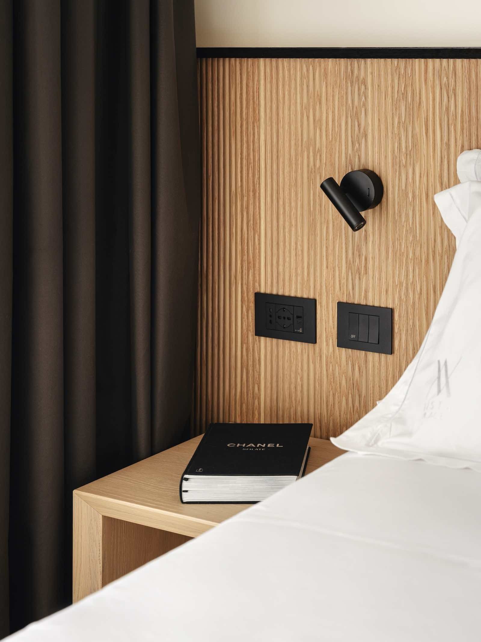 Comodino con libro, lampada e prese elettriche accanto a un letto con lenzuola bianche e una tenda marrone.