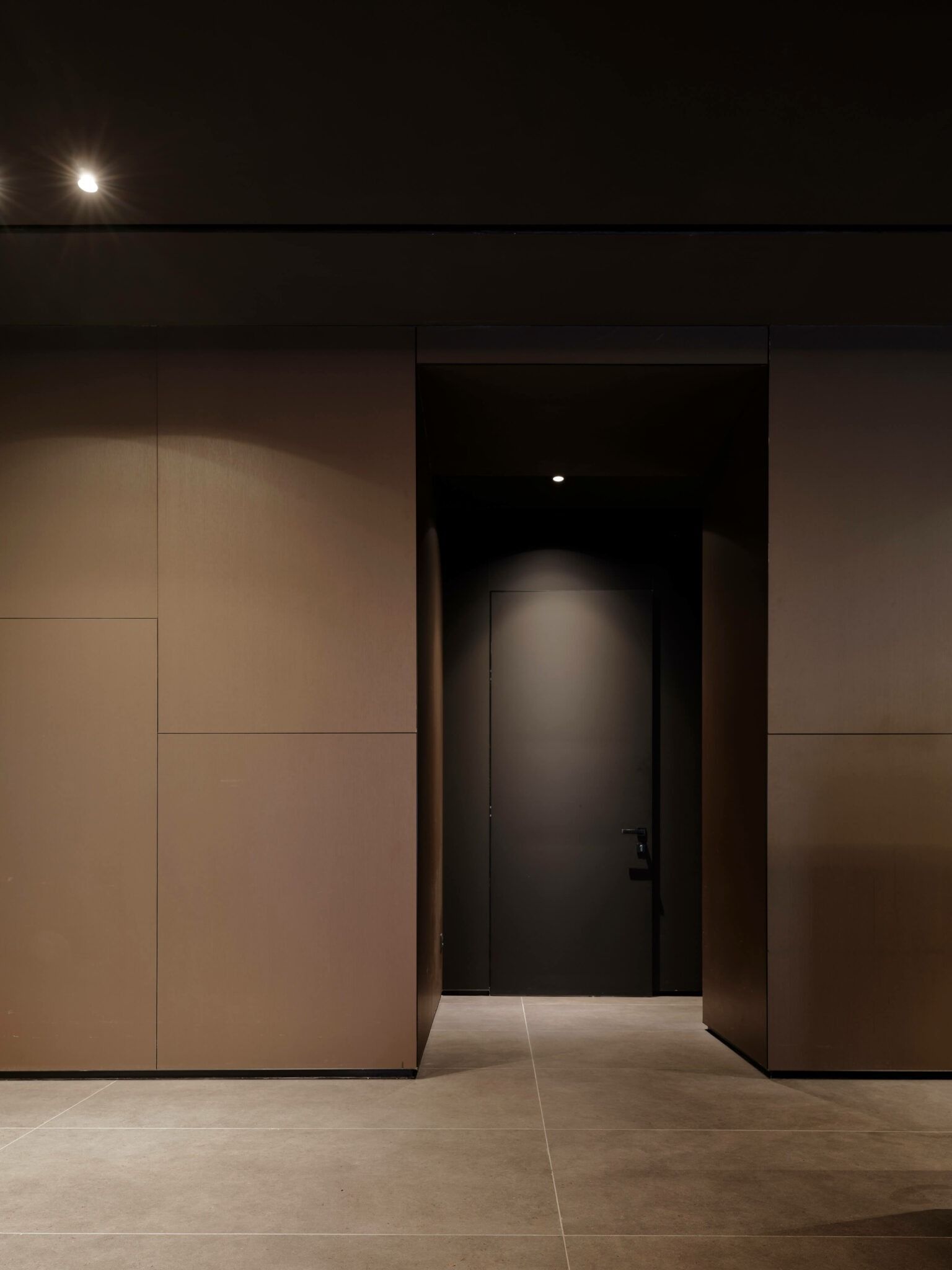 Interno buio: porta aperta su un corridoio scarsamente illuminato, fiancheggiato da pareti color cuoio e un soffitto scuro.