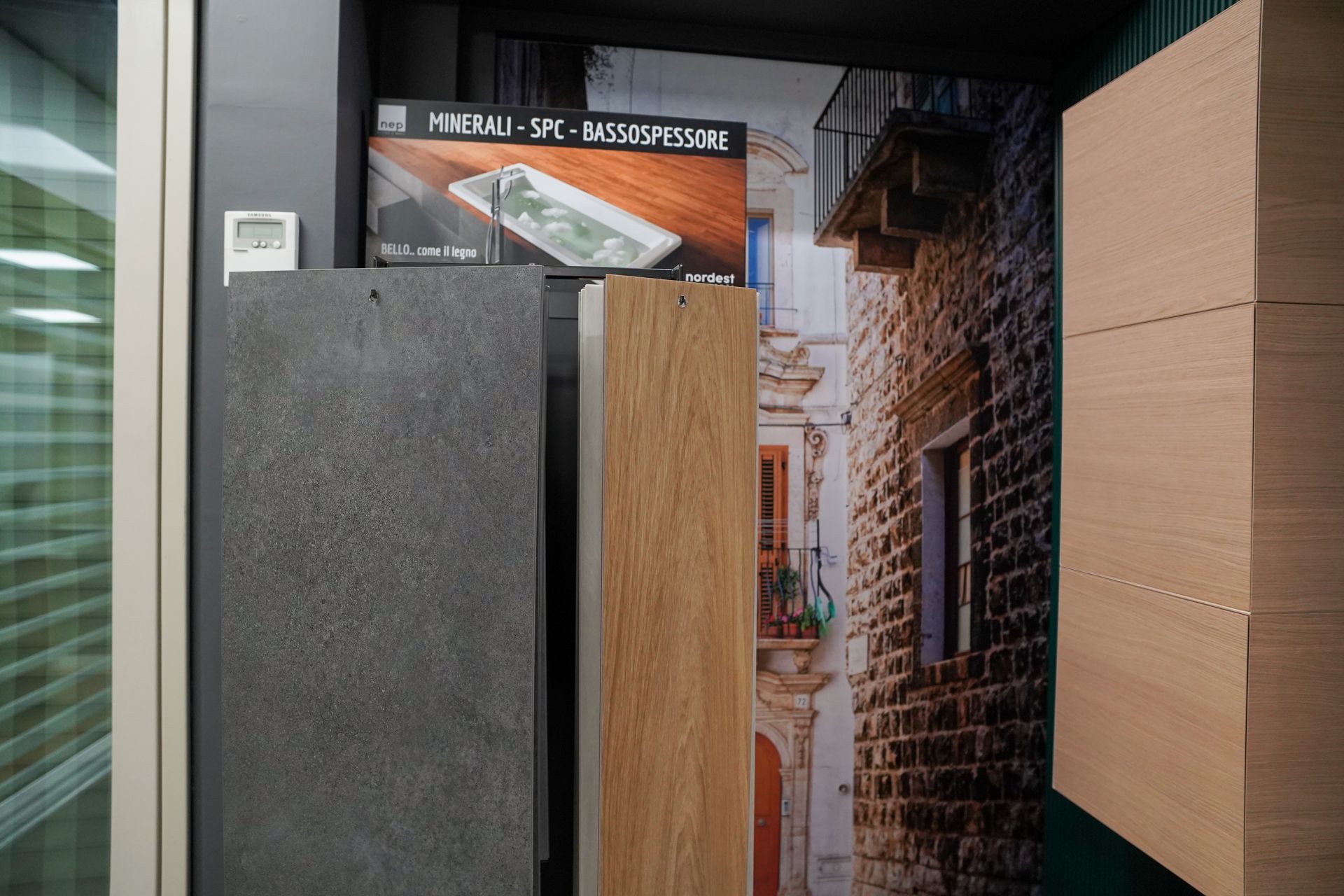 Esposizione di materiali da costruzione: piastrelle grigie, pannelli di legno, sfondo di un edificio in mattoni.