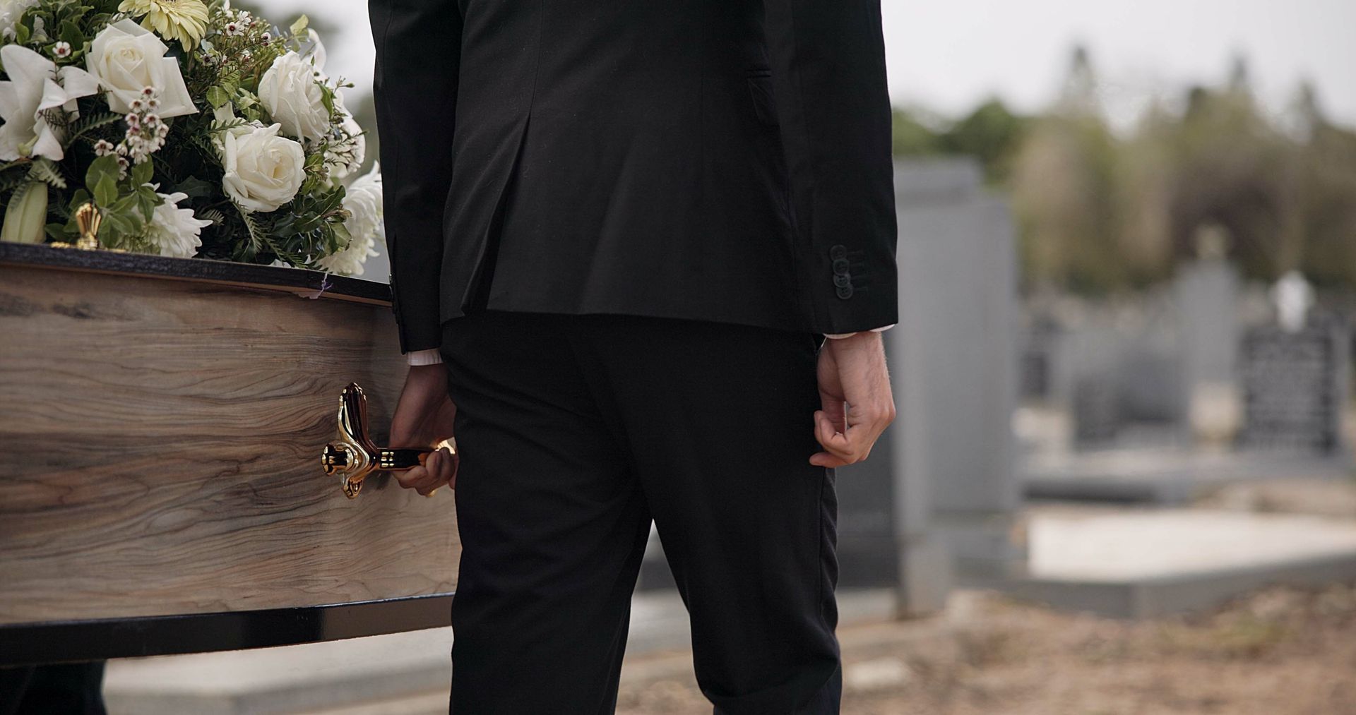 A person in a black suit stands beside a wooden casket with white flowers in a cemetery.