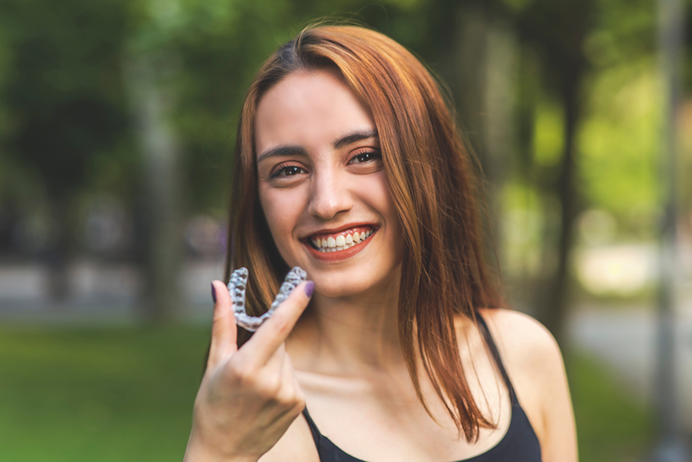 girl standing outside holding one of her Invisalign trays while smiling