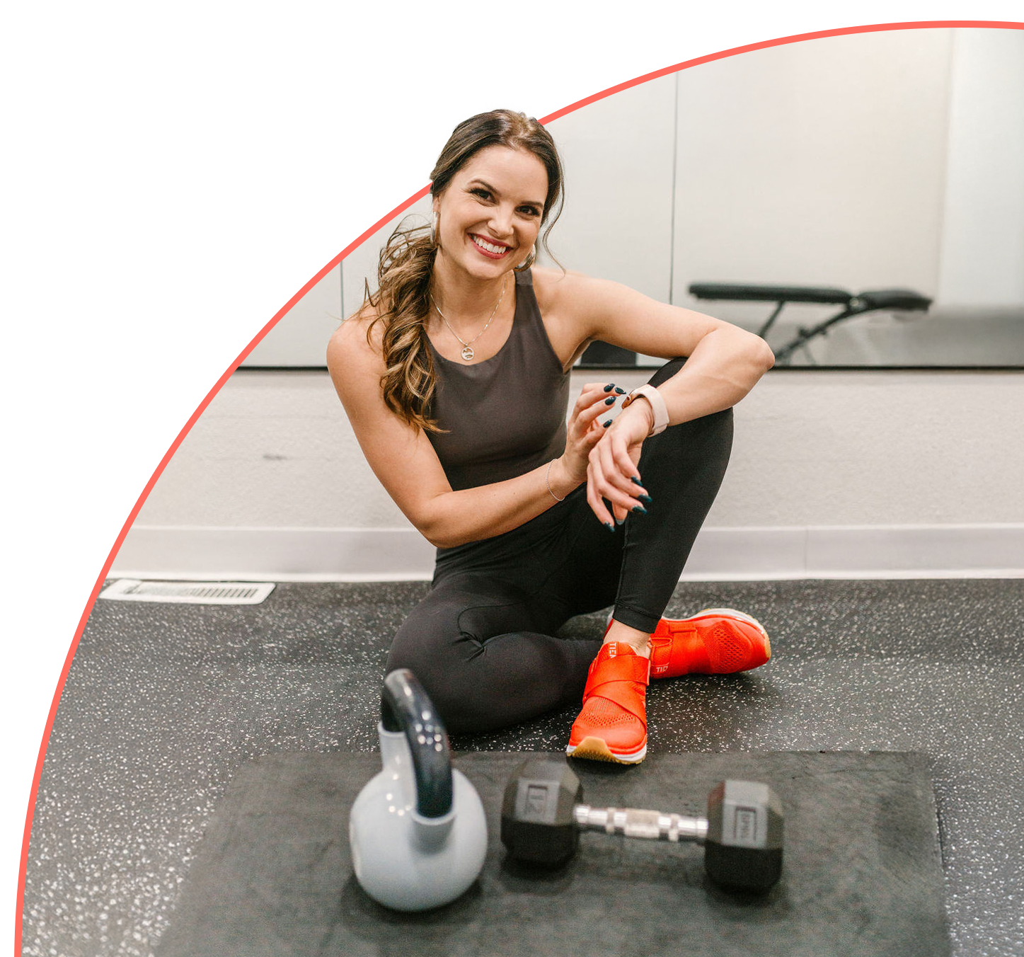 A woman is sitting on the floor in a gym next to a kettlebell and dumbbells.