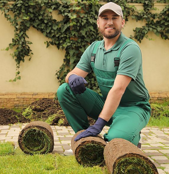 A man in green overalls is kneeling down next to rolls of grass.