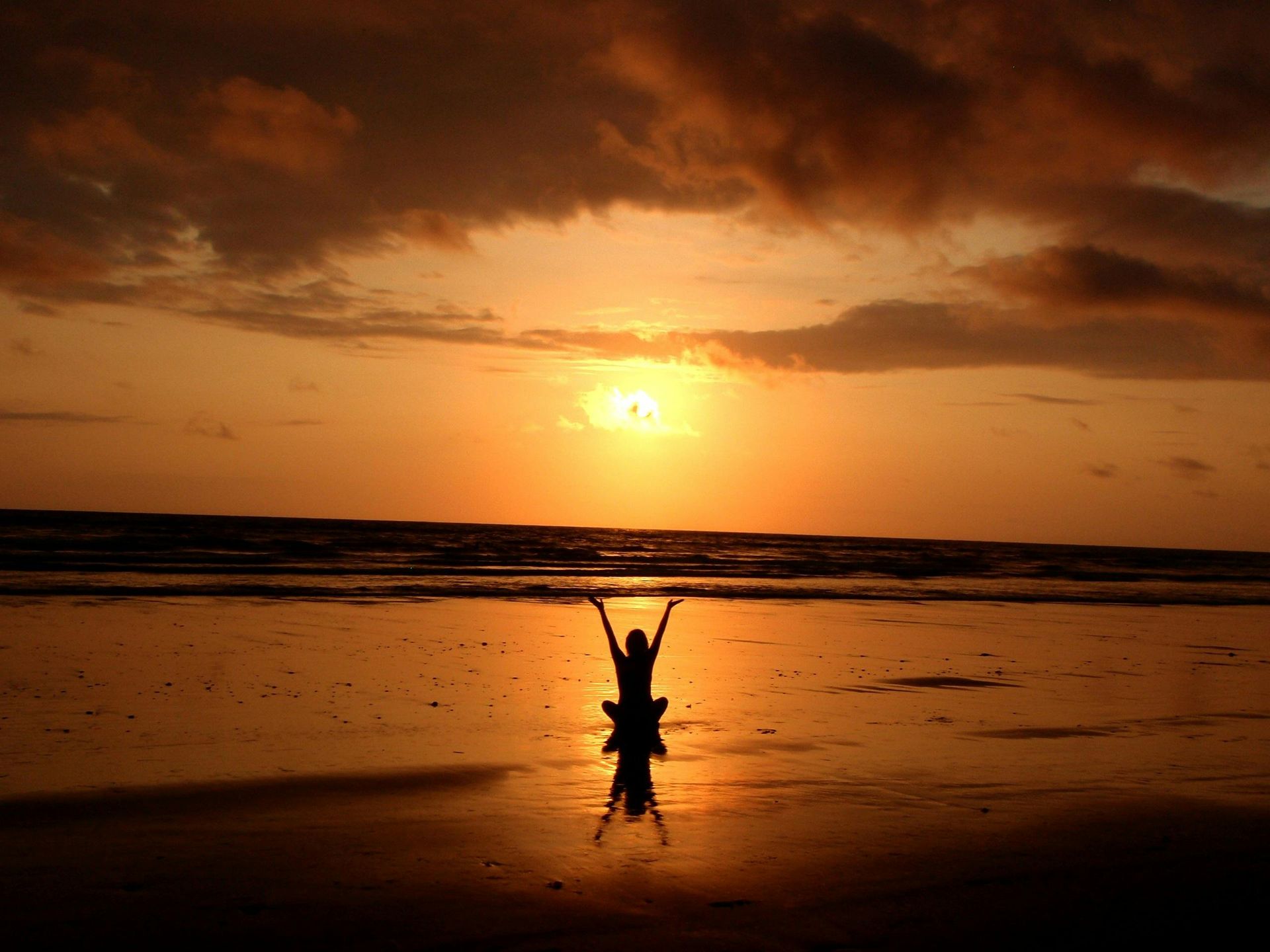 Someone sitting, crossed legs on the sea shore at sunset, with their arms lifted upward