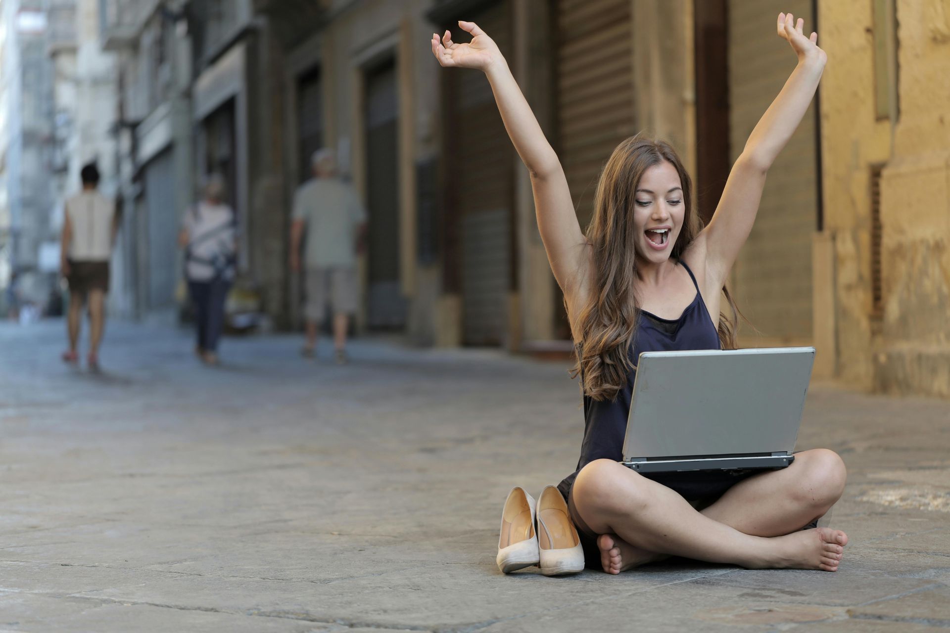 Woman sitting on the pavement with a laptop and holding her hands up in celebration