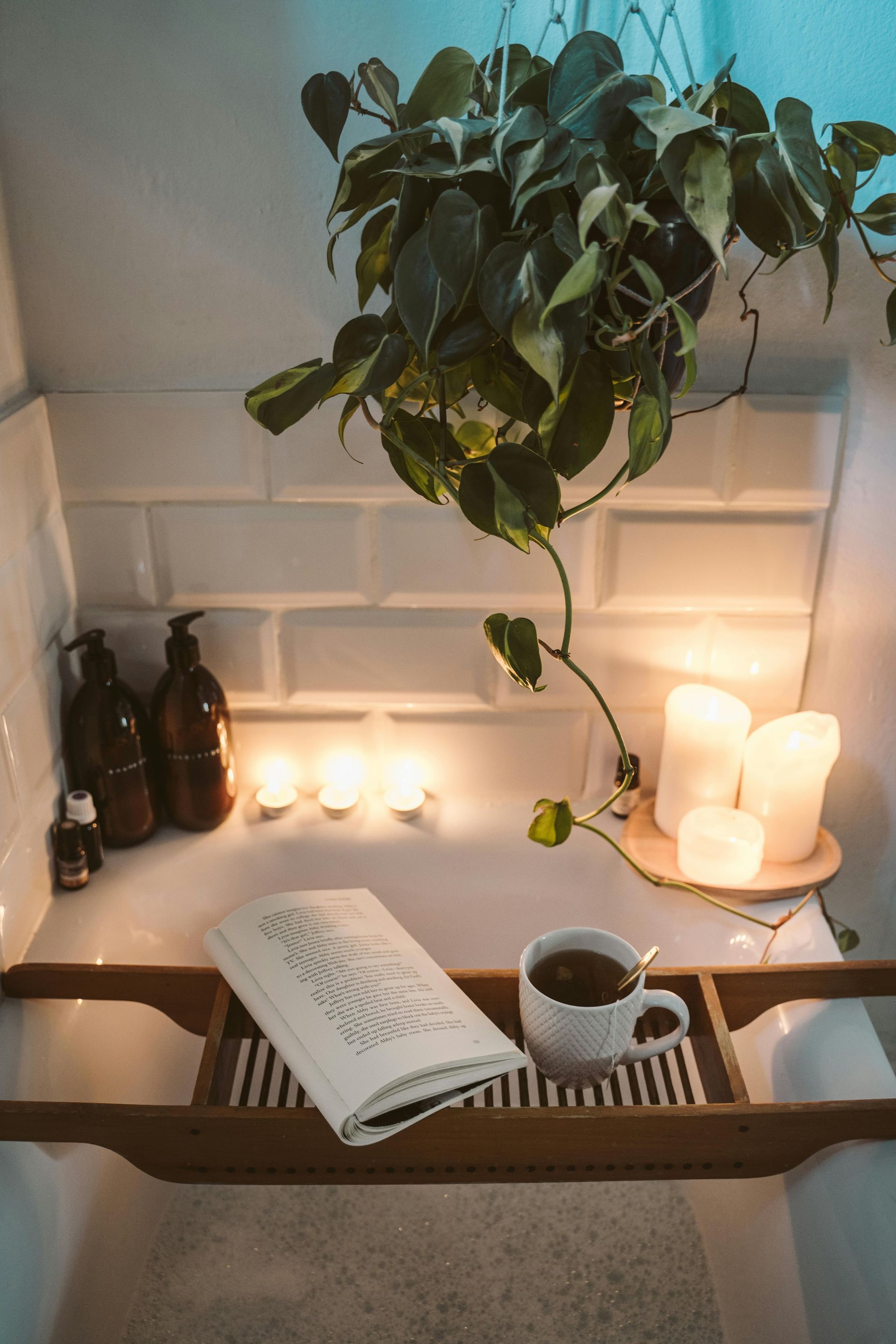 Bubble bath with candles and bottles of essential oils around the side. There's also a bathtub tray with a cup of tea and an opened book on it.