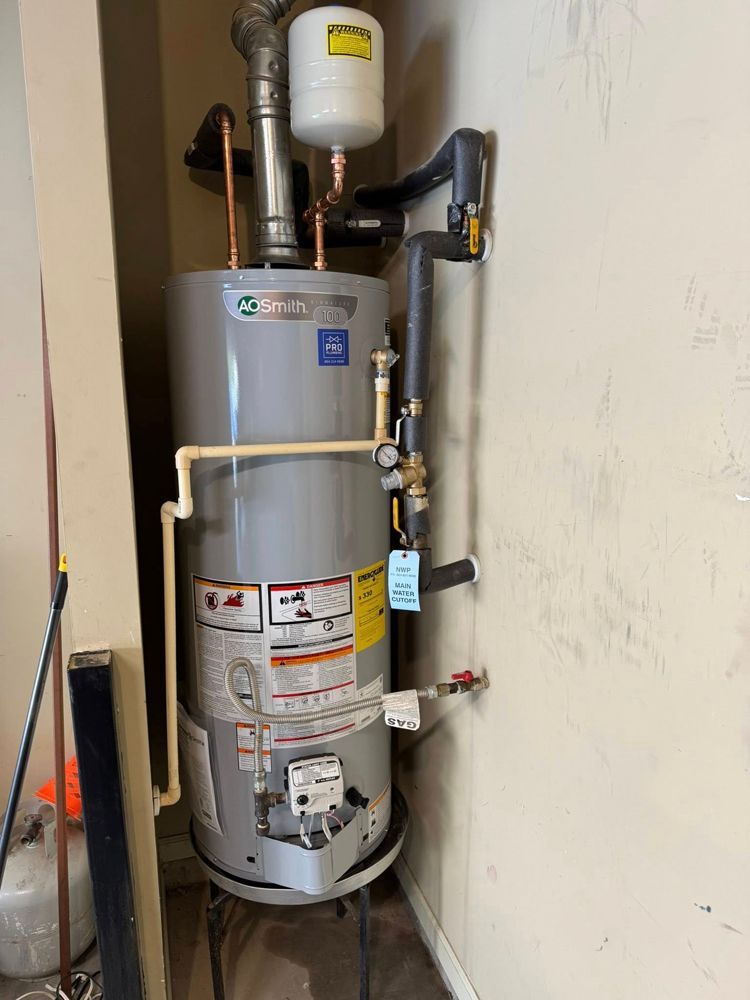 A grey water heater stands in a corner with a white expansion tank and plumbing pipes attached against a beige wall.