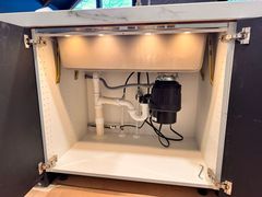 Under-sink cabinet with a white basin, garbage disposal, and plumbing pipes illuminated by installed LED lighting.