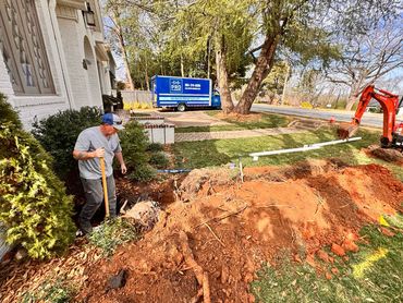 A worker digs in the dirt near a residential building, with an excavator and a blue utility truck visible in the yard.